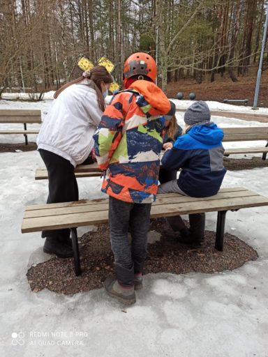 Lapset kumartuneina pöydän ääreen apuohjaajan kanssa ulkona. Maassa vielä lunta.