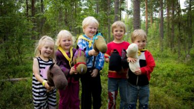 Viisi lasta nauraa metsässä keppihevosten selässä.