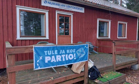 Punainen ppuurakennus, jossa kyltti Nummelan lentokeskus. Terassille sidottu banderolli, jossa lukee Tule ja koe partio.